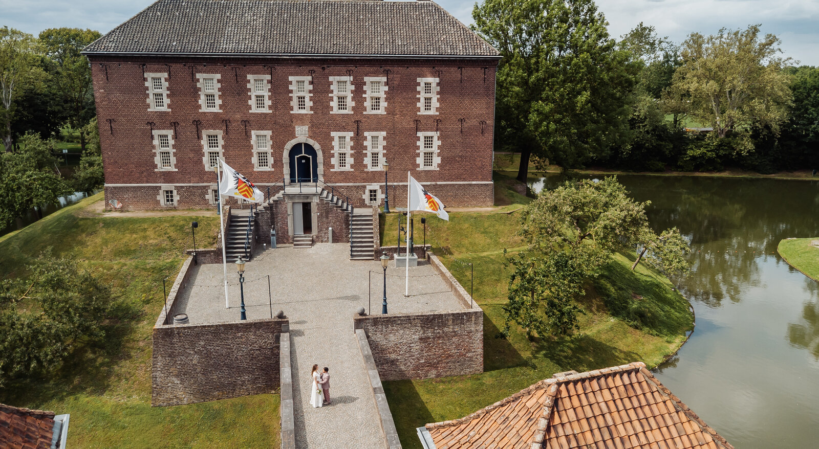 Luchtfoto van bruidspaar Sofie en Jaya dat over de toegangsbrug naar Kasteel Limbricht loopt, met de slotgracht op de achtergrond