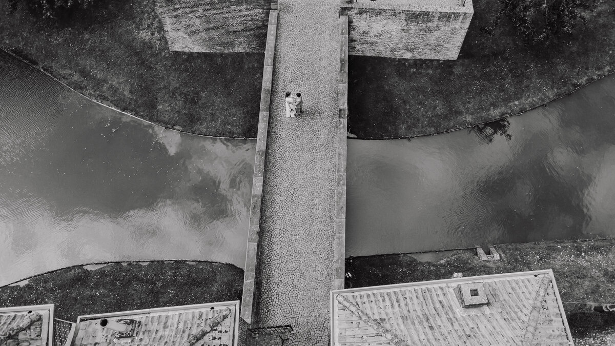 Dramatische zwart-wit dronefoto van bruidspaar op de brug van Kasteel Limbricht, omringd door de slotgracht